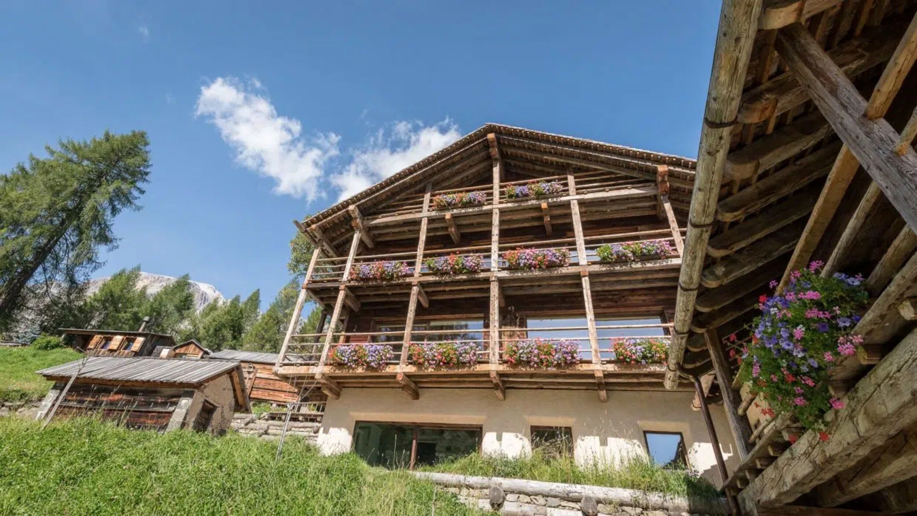 A wooden chalet with multiple balconies, each adorned with colorful flowers, set in a mountainous area.