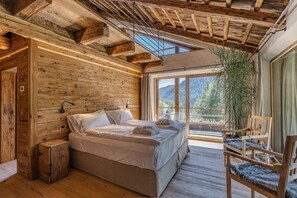 A wooden cabin bedroom with a bed, a chair, and a large window overlooking a mountainous landscape.