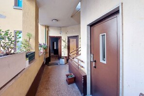 A narrow corridor with a brown door on the right, a white door in the background, and potted plants along the left wall.