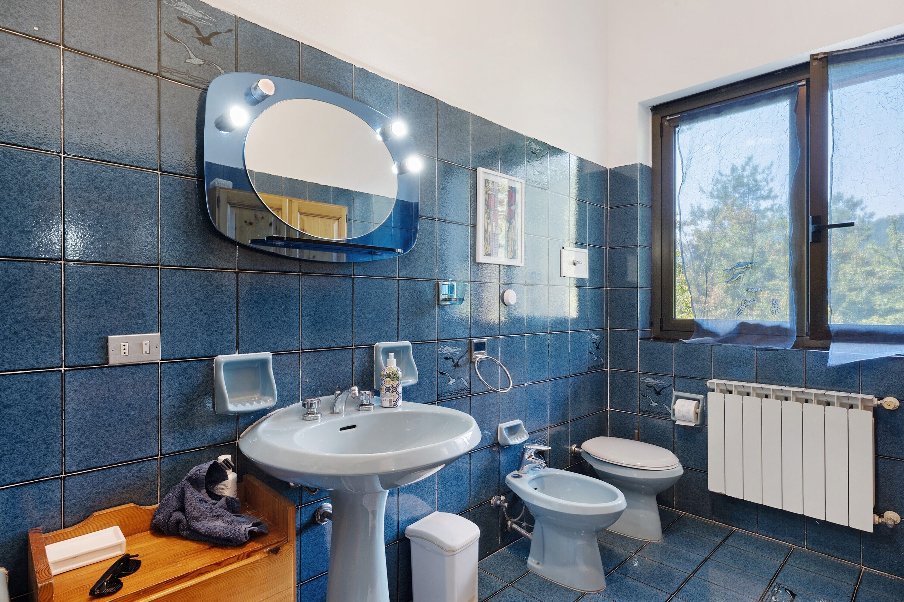 Ein Badezimmer mit Waschbecken, Spiegel, Toilette, Bidet und einem Fenster mit Blick auf Bäume.