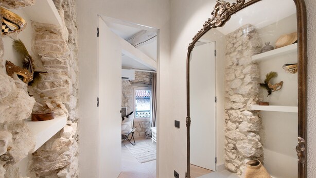 A hallway with stone walls, a mirror, and a door leading to a room with a bed and a window.