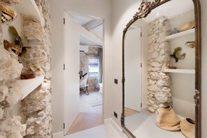 Un pasillo con paredes de piedra, un espejo y una puerta que da a una habitación con cama y ventana.