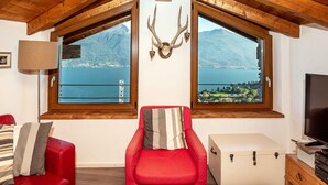 A room with a red sofa, a wooden chest, and a TV. There are two windows with a mountain view. - Holiday Home 'Casa Bianca' with Lake View, Private Terrace and Wi-Fi (Saliana (Pianello del Lario))