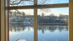 Une vue à travers une fenêtre sur un lac paisible, avec un quai, entouré d’arbres et de maisons.