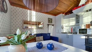 Un salon avec un canapé bleu, une table blanche ornée de deux vases bleus et une plante en pot. On aperçoit, au fond, une cuisine avec des meubles blancs et un four à micro-ondes.