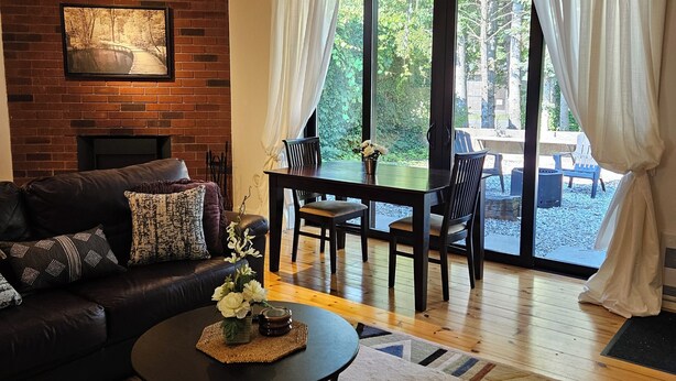 Un salon avec un canapé en cuir, une table basse et une cheminée. On y trouve également une table à manger avec des chaises, et de grandes fenêtres offrent une vue sur l’extérieur.