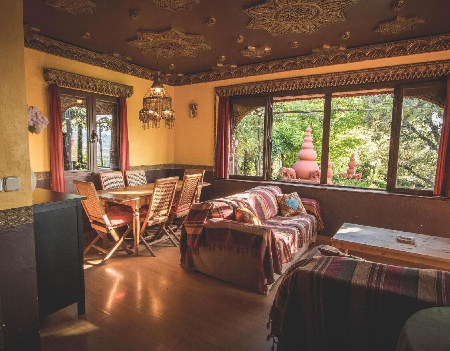 A room with a dining table, chairs, a sofa, and a window with a view of greenery and a pink pagoda.