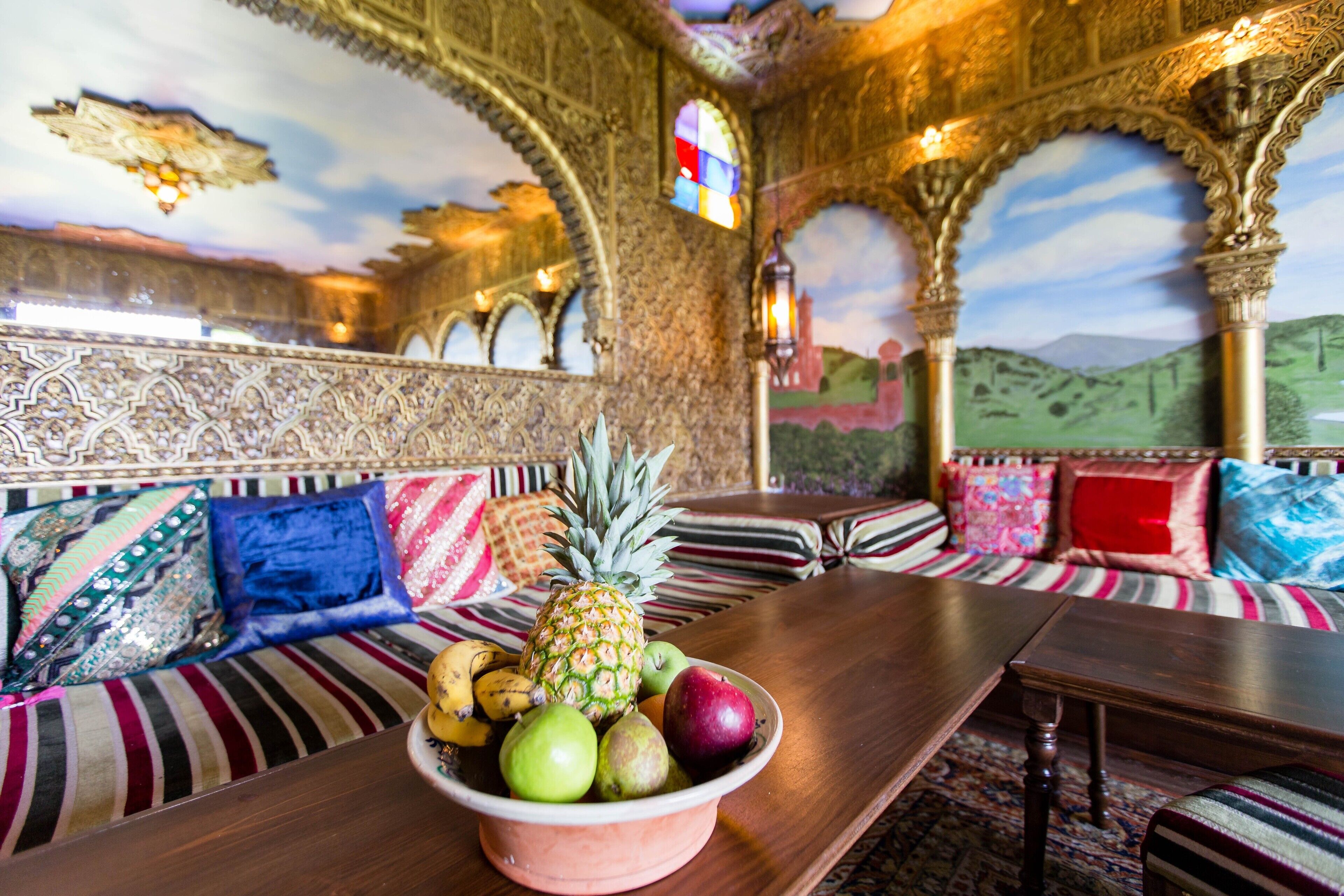 Una habitación con una mesa de madera, un frutero con fruta, un sofá con cojines estampados y decoración de pared ornamentada.