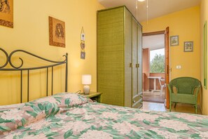Une chambre à coucher avec un lit, un fauteuil vert, une petite table avec une lampe et une porte-fenêtre donnant sur un balcon.