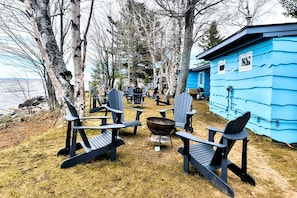 Eine blaue Holzhütte mit Adirondack-Stühlen, die um eine Feuerstelle angeordnet sind, vor dem Hintergrund von Bäumen und einem Gewässer.