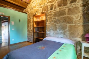 Une pièce avec un mur en pierre, un lit recouvert d’une couvre-lit à motifs floraux, une étagère en bois et une table de chevet blanche avec une lampe.