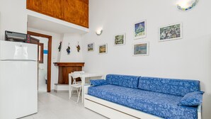 A compact kitchen with a white refrigerator, a wooden cabinet, a white dining table with chairs, and a blue sofa with patterned cushions.
