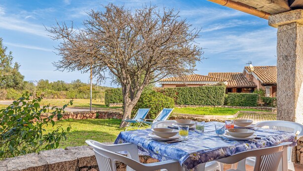 An outdoor dining area with a table set for four, surrounded by white plastic chairs, a stone wall, and a view of a house with a tiled roof.