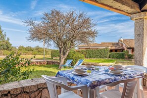 An outdoor dining area with a table set for four, surrounded by white plastic chairs, a stone wall, and a view of a house with a tiled roof. - Holiday Home 'Villetta A 800mt Dall'Isuledda' with Shared Terrace (san teodoro)