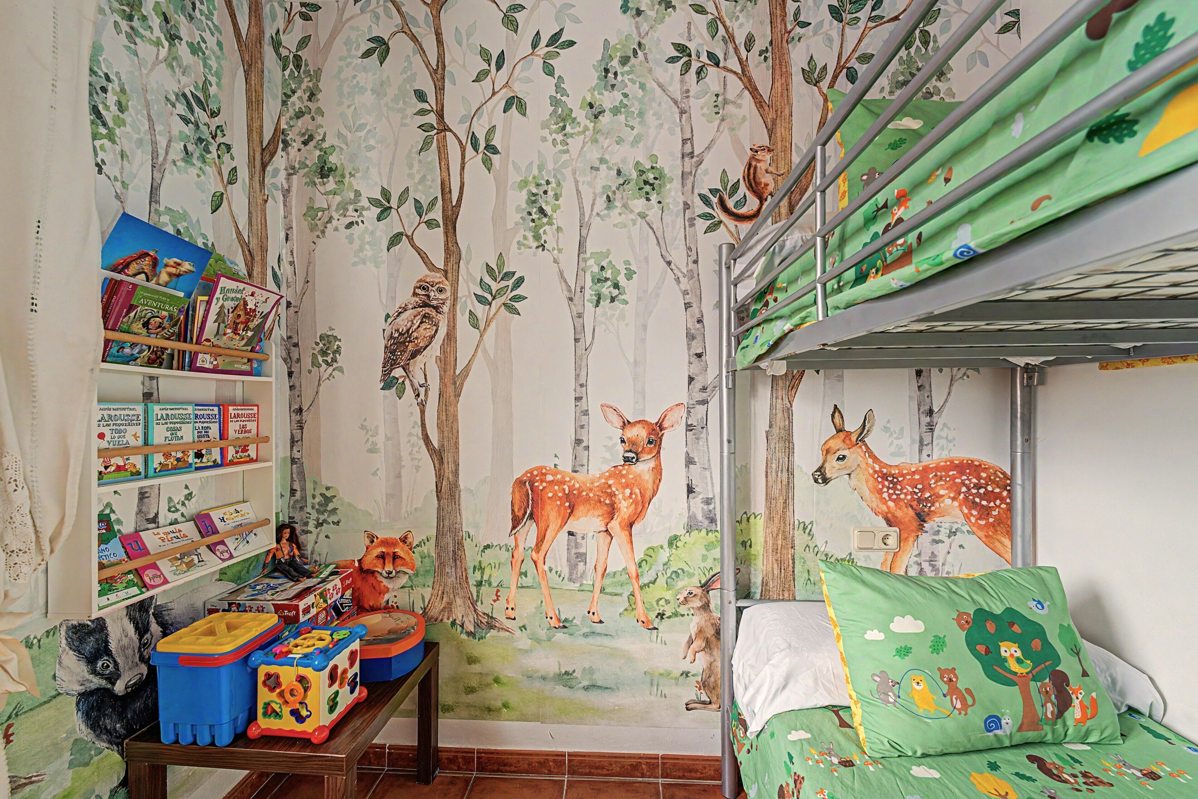 Habitación infantil con mural temático de bosque, estantería, mesa de juegos y literas con edredón verde.