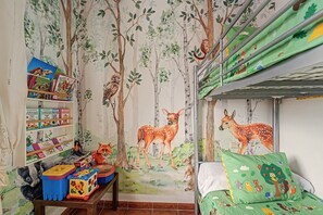 Une chambre d’enfant avec une fresque murale sur le thème de la forêt, une bibliothèque, une table pour jouer et un lit superposé avec une couverture verte.
