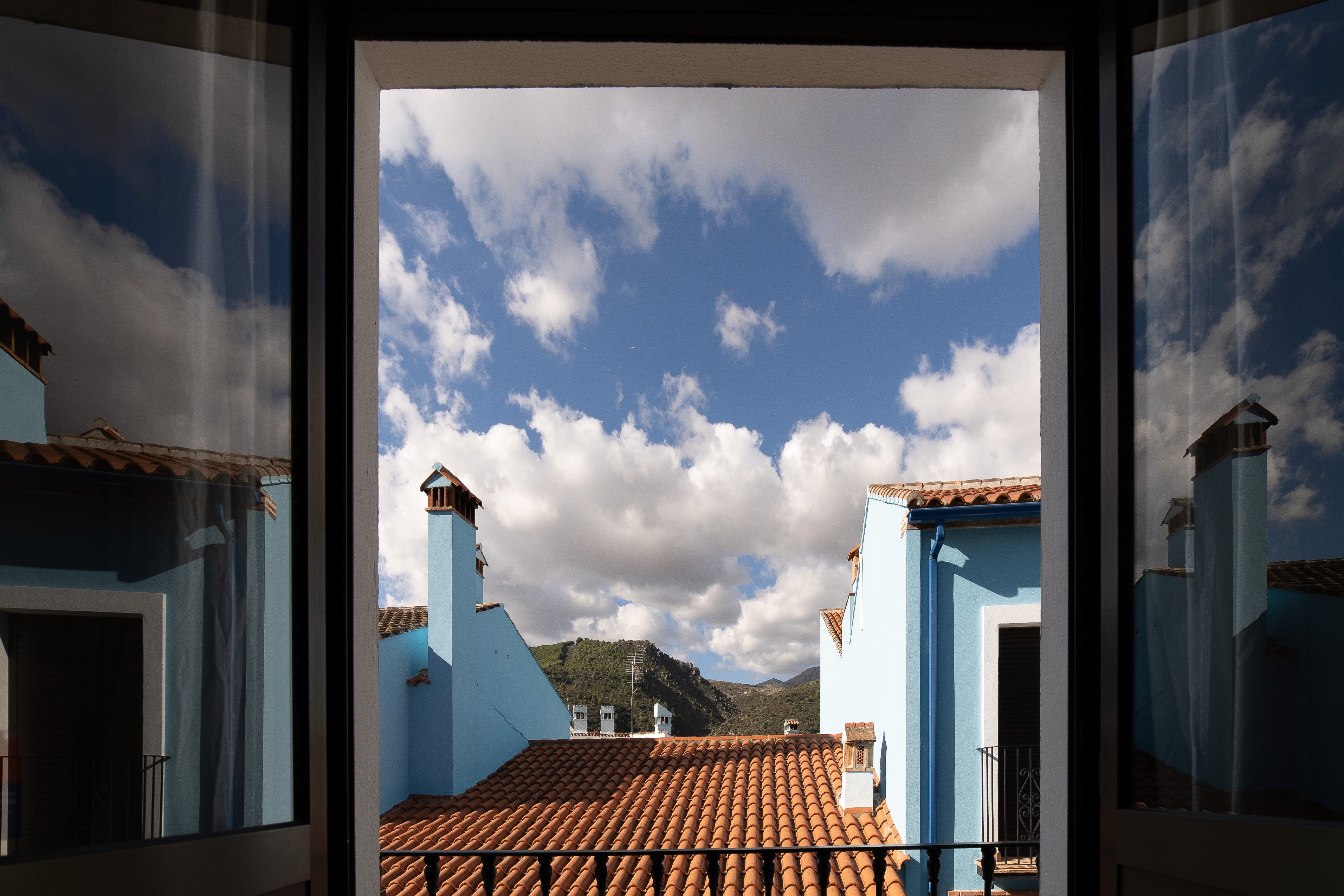 A view through a window of a building with a tiled roof and blue walls, with a mountain and cloudy sky in the background.