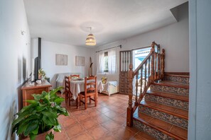 Un espace repas avec une table et des chaises, un escalier avec des balustrades en bois et une vue sur l’extérieur par une porte.