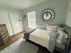 Une chambre à coucher avec un lit, une horloge murale, une fenêtre avec des stores, une lampe sur pied et une commode.