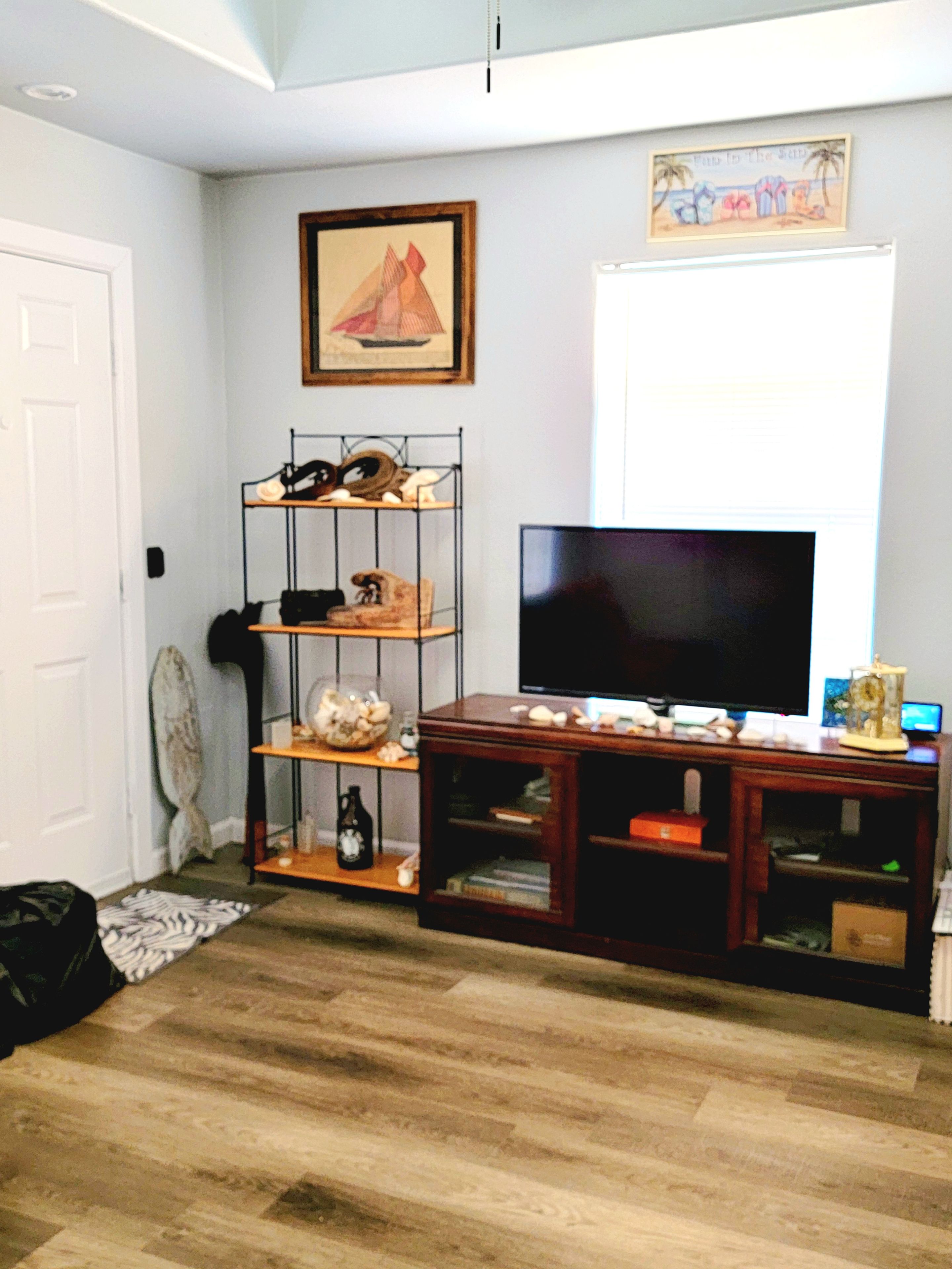 Una sala de estar con piso de madera, un televisor sobre un mueble de madera, una estantería con varios objetos y una puerta.