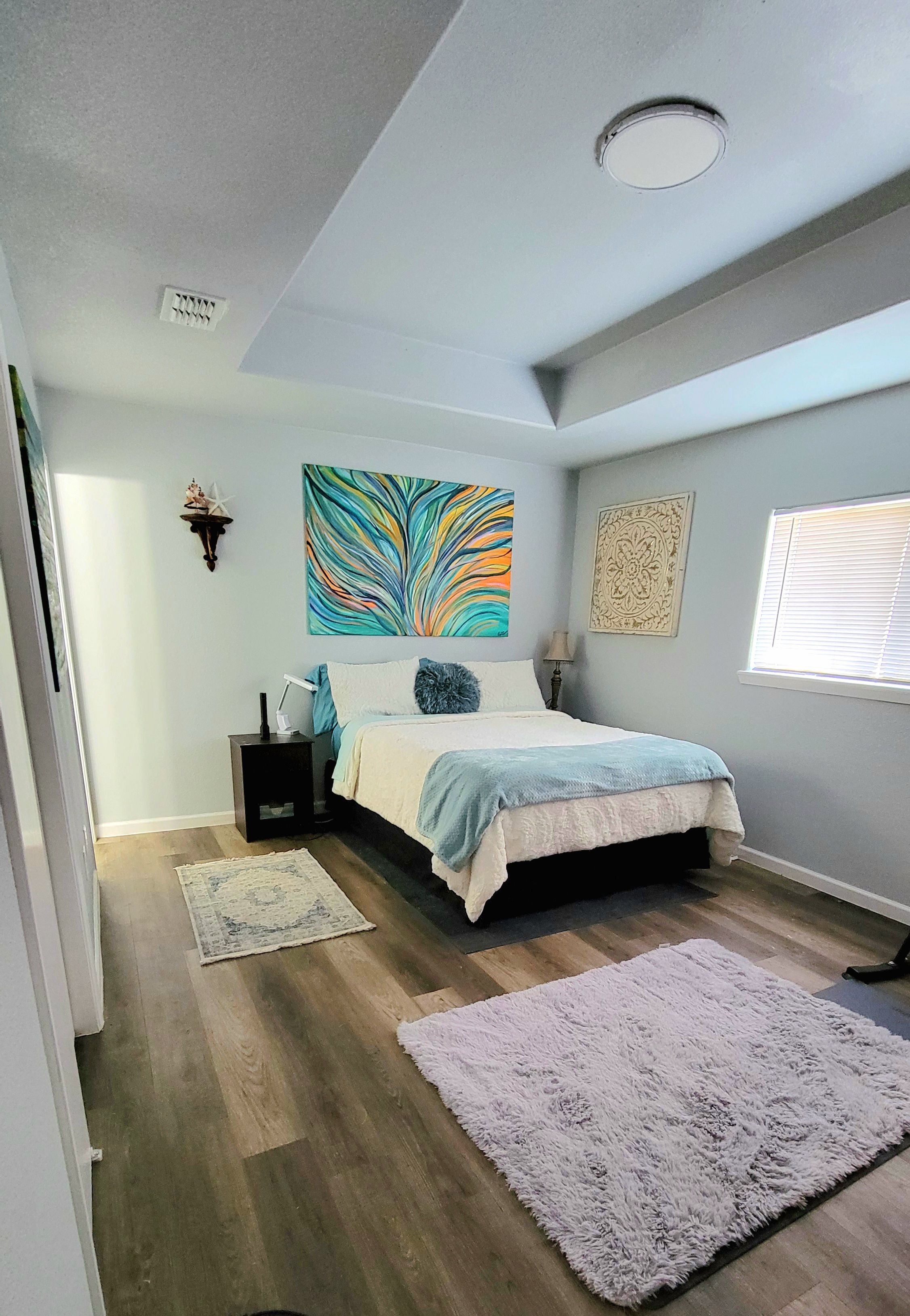 A bedroom with a bed, a nightstand, a wall-mounted lamp, a decorative star, a colorful abstract painting, a patterned rug, and a window with blinds.