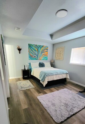 Een slaapkamer met een bed, nachtkastje, een wandlamp, een decoratieve ster, een kleurrijk abstract schilderij, een tapijt met een patroon, en een raam met jaloezieën.