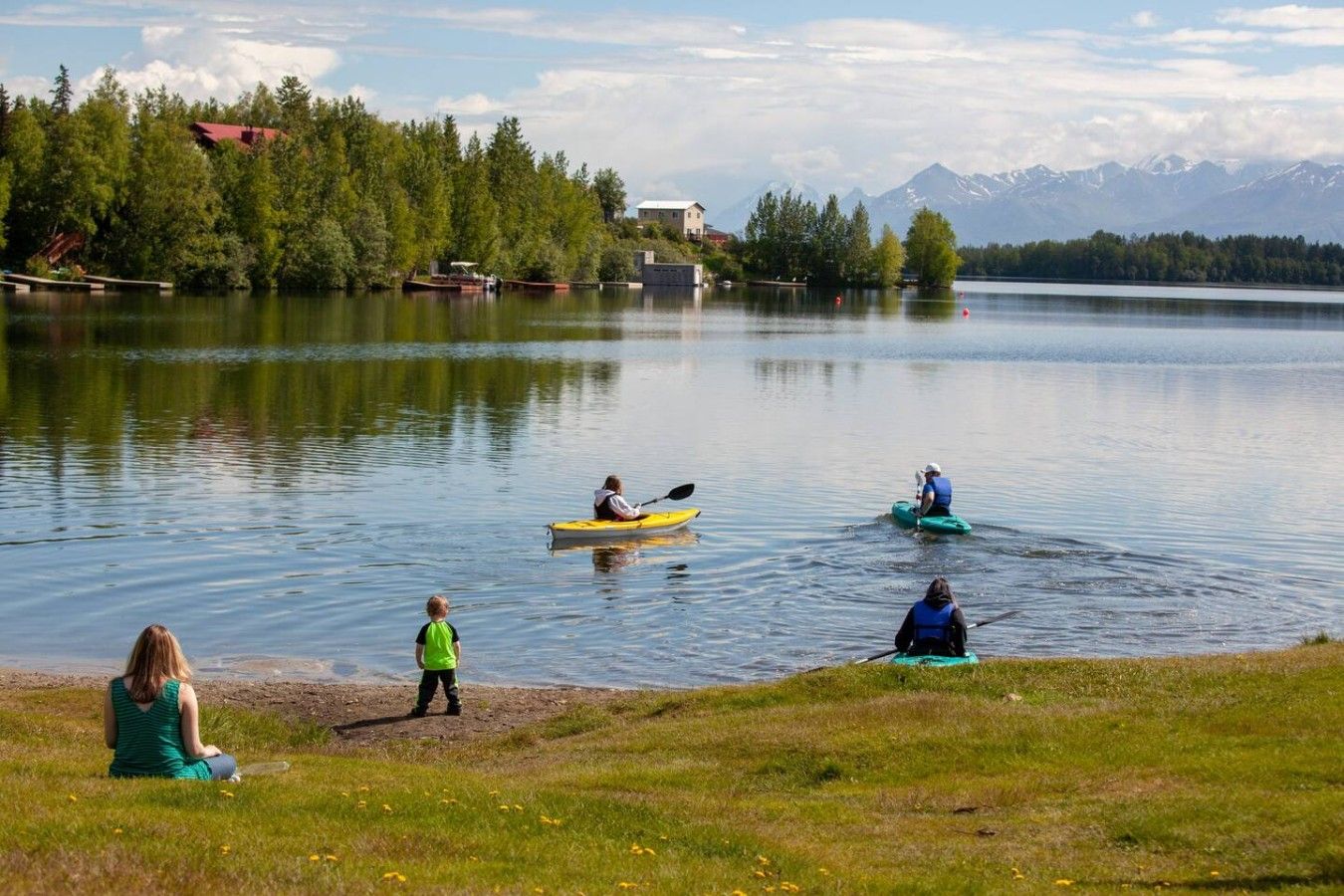 Un lago tranquilo con kayaks, una orilla cubierta de césped y un área boscosa con edificios al fondo.