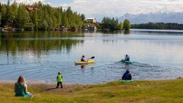 Un lago tranquilo con kayaks, una orilla cubierta de césped y un área boscosa con edificios al fondo.