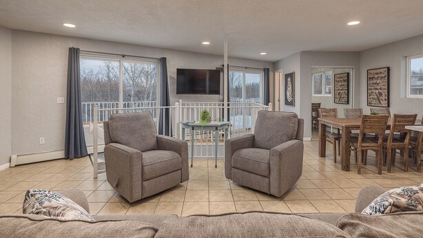 A modern living room with grey sofas, a dining table, and a balcony with a TV. - 2-Story Lakeside Oasis w/ Private Dock, Kayaks & Fire Pit (Wasilla)