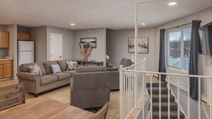 A modern living room with a sectional sofa, a dining table, and a staircase leading to a balcony. - 2-Story Lakeside Oasis w/ Private Dock, Kayaks & Fire Pit (Wasilla)