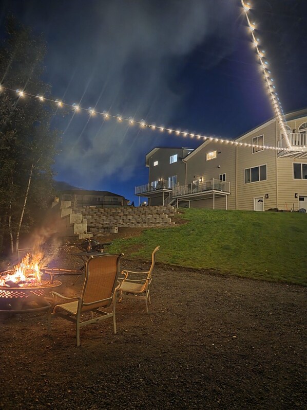 A two-story building with a fire pit and chairs in the foreground, and string lights above. - 2-Story Lakeside Oasis w/ Private Dock, Kayaks (Wasilla)