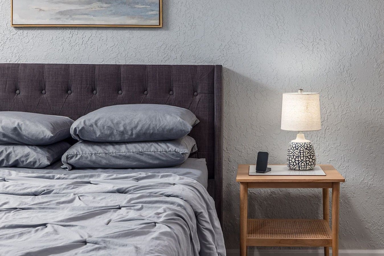 A neatly made bed with grey bedding and pillows, a bedside table with a lamp, and a textured wall.