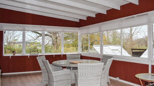 A sunroom with a round table and wicker chairs, large windows, and a view of trees and a greenhouse.