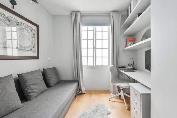 Ein modernes Schlafzimmer mit einem grauen Sofa, einem Schreibtisch mit Stuhl, einem Bücherregal und einem großen Fenster mit Vorhängen.
