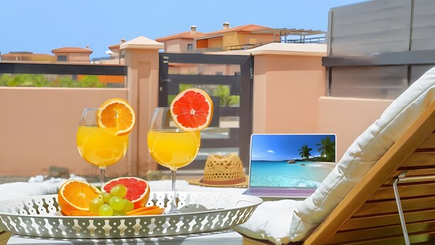 A sunlit outdoor seating area with a laptop displaying a beach view, a fruit tray, and a straw in a glass. - Vacation 'Bonita Lajares 3' with Shared Pool, Private Terrace and Wi-Fi (Lajares)