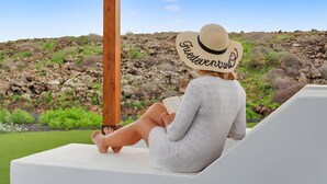 A person sitting on a white lounge chair reading a book with a hat labeled 'Flecheventura' against a rocky landscape backdrop.