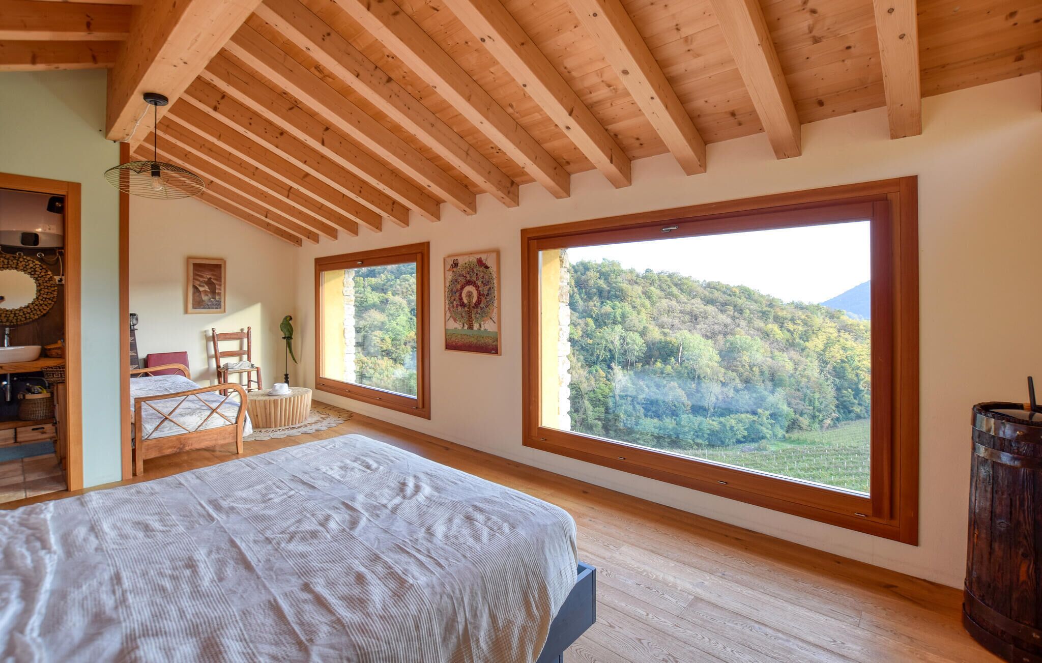 Una camera da letto con una grande finestra, travi in legno, un letto, una sedia, un tavolino e un lampadario.