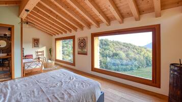 Una camera da letto con una grande finestra, travi in legno, un letto, una sedia, un tavolino e un lampadario.