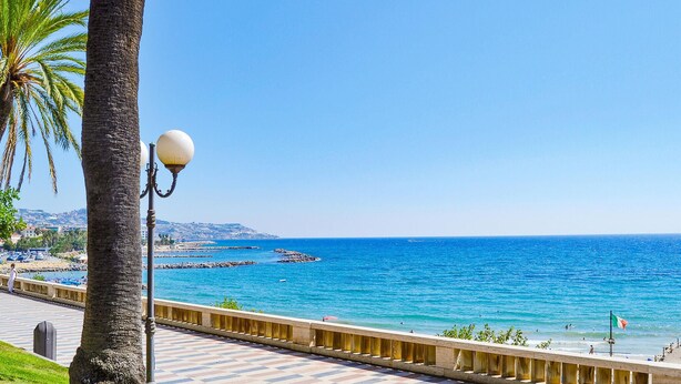 Eine Küstenpromenade mit freiem Blick auf das Meer, Palmen und einer Laterne.