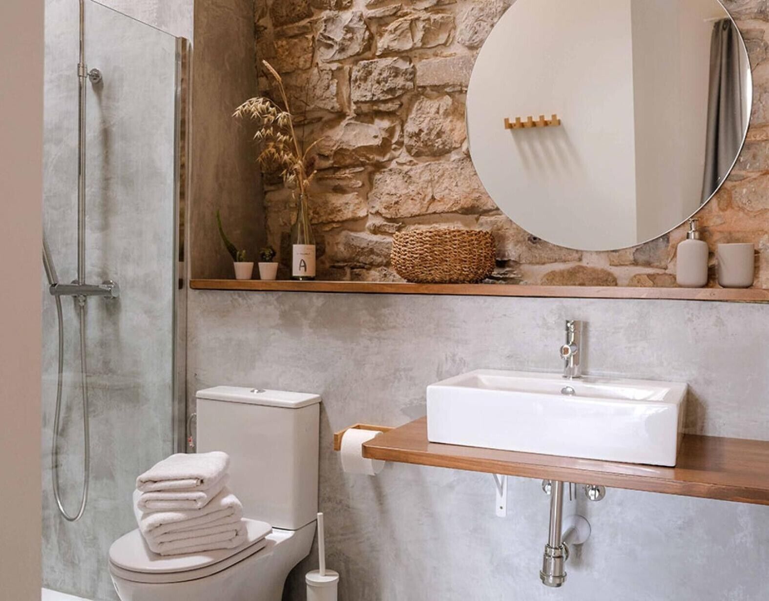 A bathroom with a stone wall, a round mirror, a white sink, a toilet, and a shower.