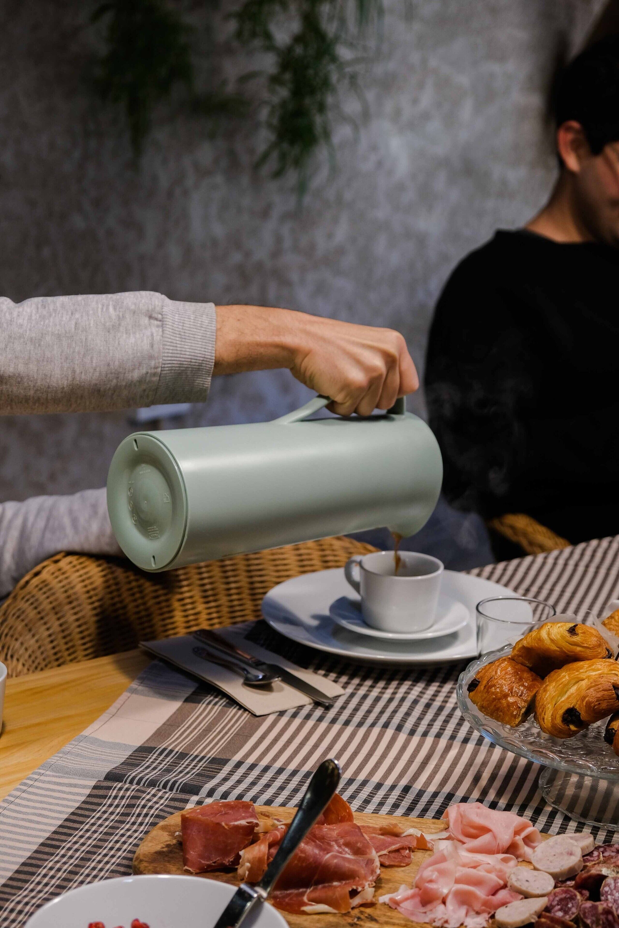 Une table mise pour le petit-déjeuner, avec une cafetière, des croissants et diverses sortes de charcuterie, le tout posé sur une nappe à carreaux.