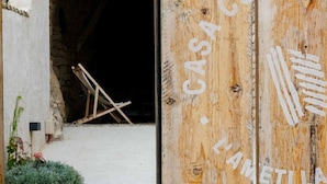 Een houten deur, waarop 'CASA COMAL' en 'L'AMETTLA' in hout gegraveerd staan, die leidt naar een interieur met stenen muren.