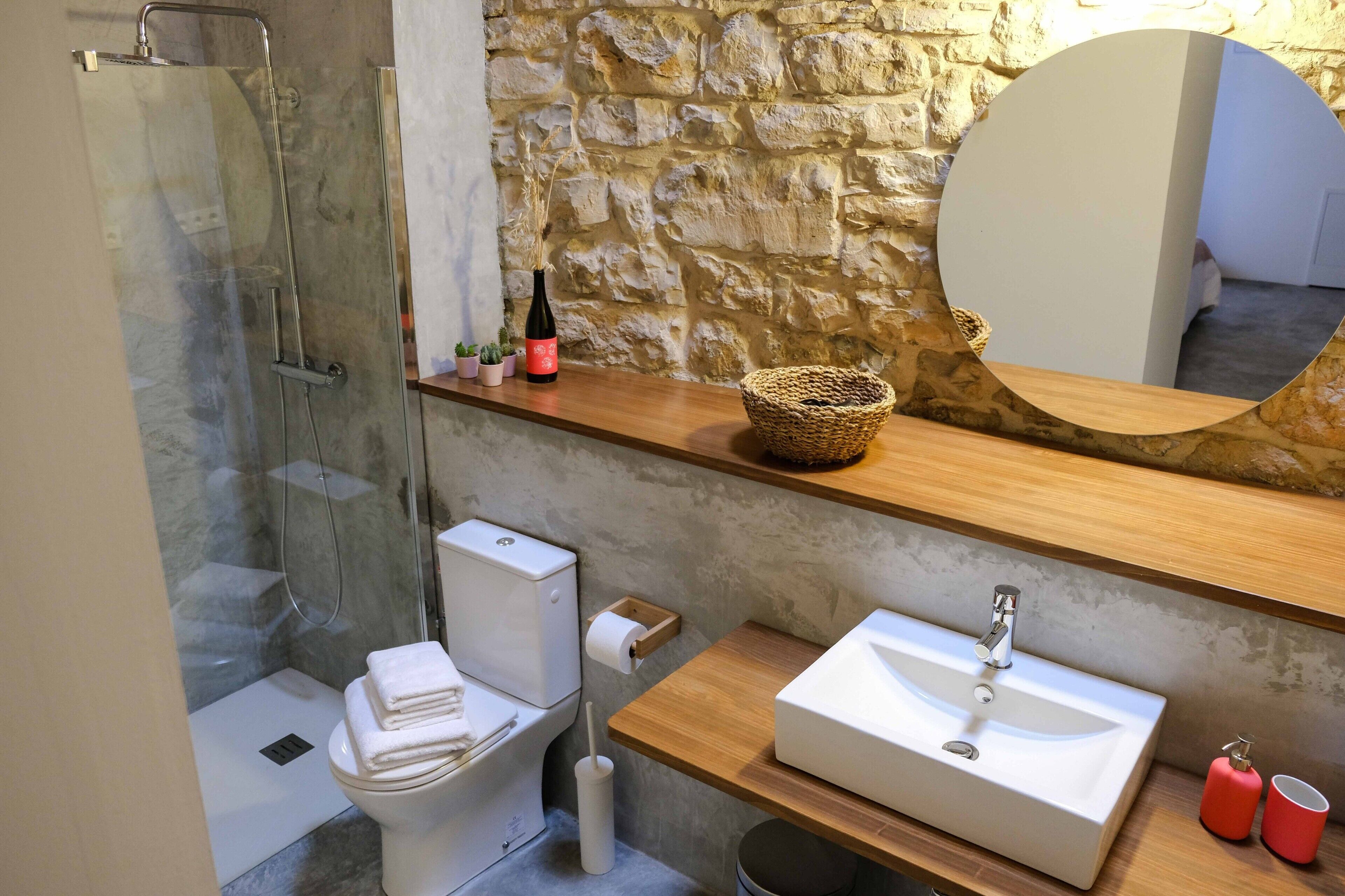 Une salle de bain moderne avec une douche à l’italienne en verre, des toilettes, un lavabo avec un miroir au-dessus et un mur en pierre.