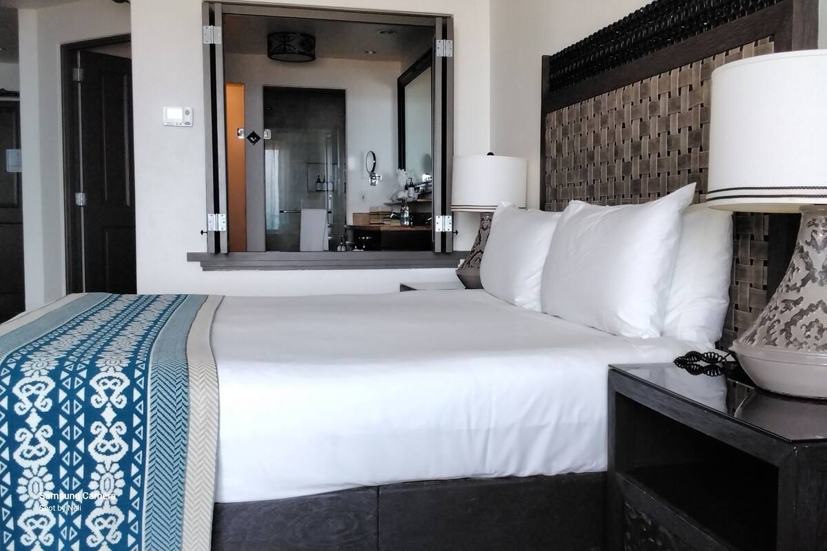 A hotel room with a large bed, a bedside table with a lamp, a mirror, and a view of the bathroom.