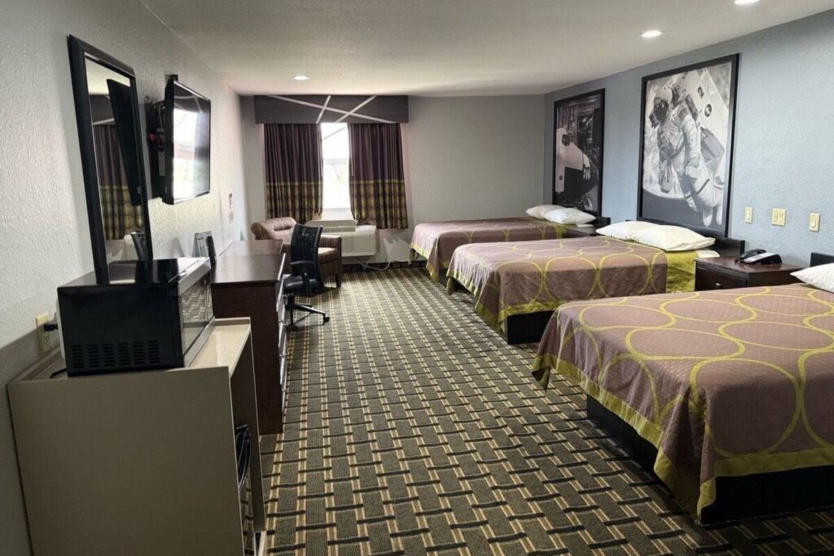 Habitación de hotel con tres camas, un escritorio, una silla, un televisor y un cartel grande en la pared.