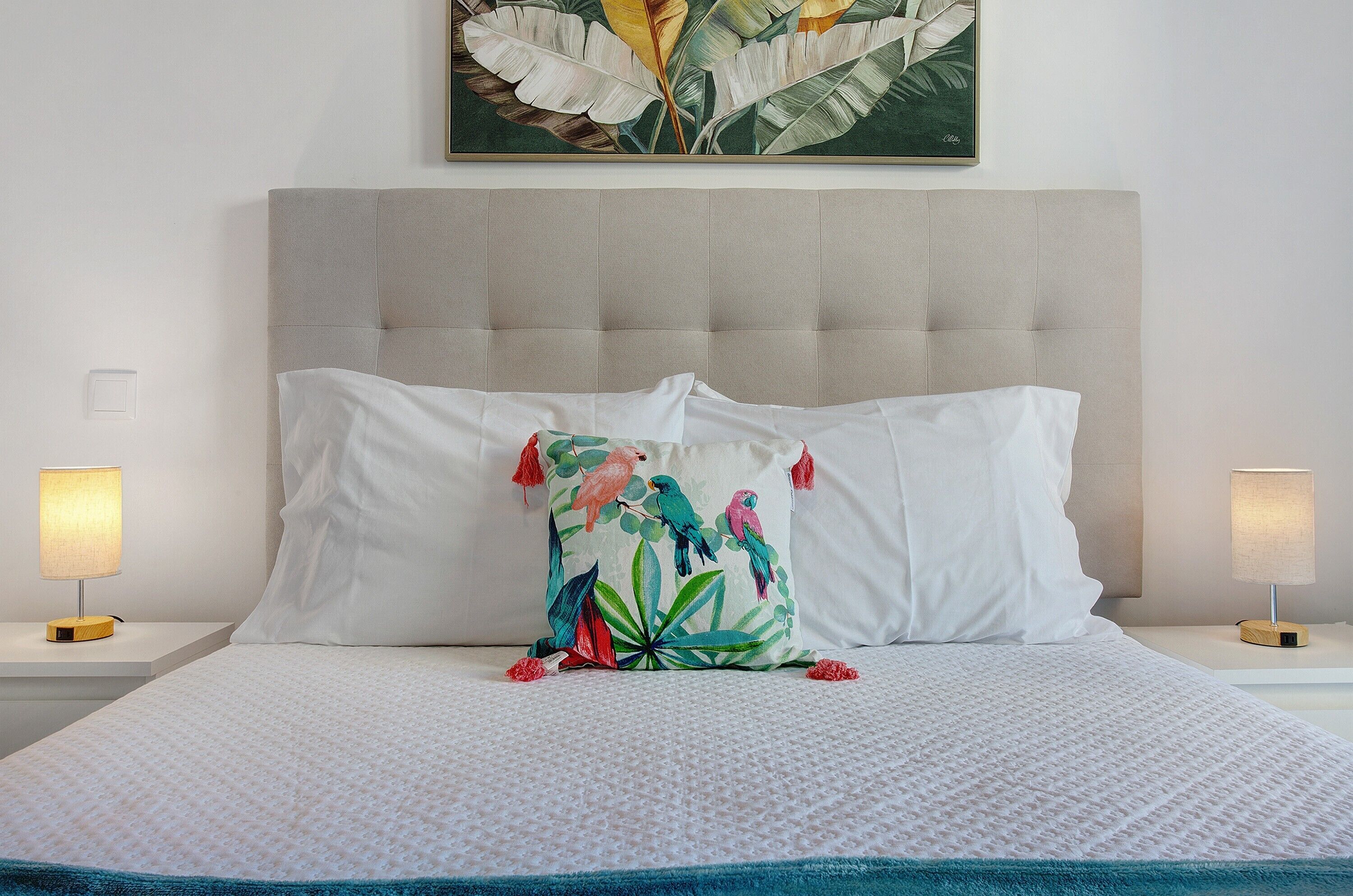 Een net opgemaakt bed met een witte dekbedovertrek, een decoratief kussen met vogels en twee bedlampen op witte nachtkastjes.