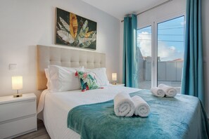 Une chambre d’hôtel avec un grand lit, une table de chevet avec une lampe, une fenêtre avec des rideaux et un tableau représentant des feuilles tropicales accroché au mur.