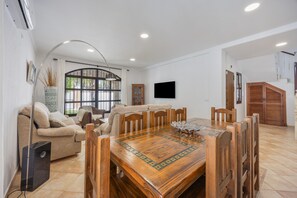 A dining area with a wooden table and chairs, a sofa, a TV mounted on the wall, and a window with a view.