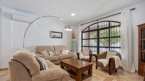 Una sala de estar con sofá, sillones, una mesita de centro, un ventanal con cortinas y un aire acondicionado instalado en la pared.