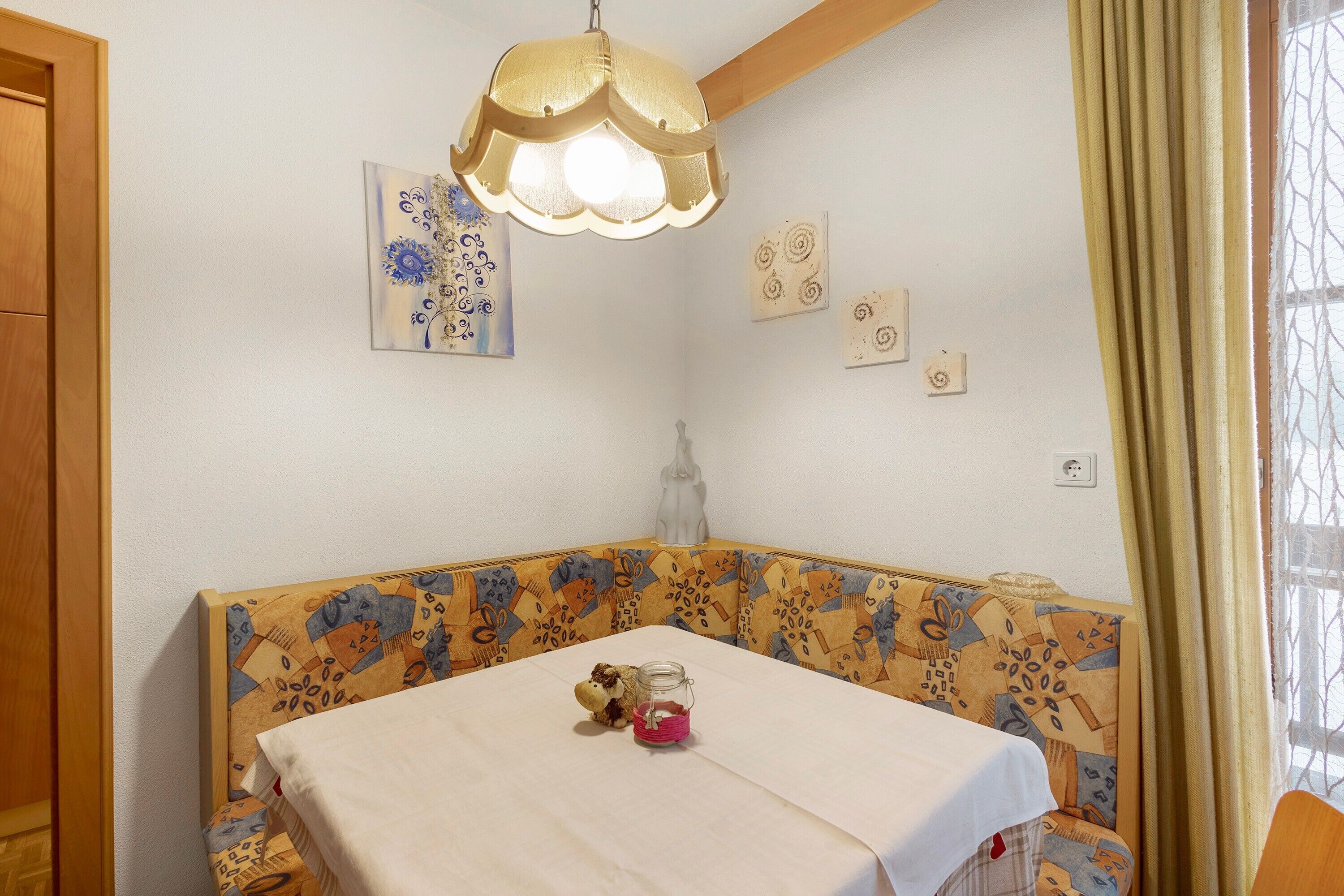 A corner room with a patterned bench, a table with a white tablecloth, a decorative light fixture, and wall art.